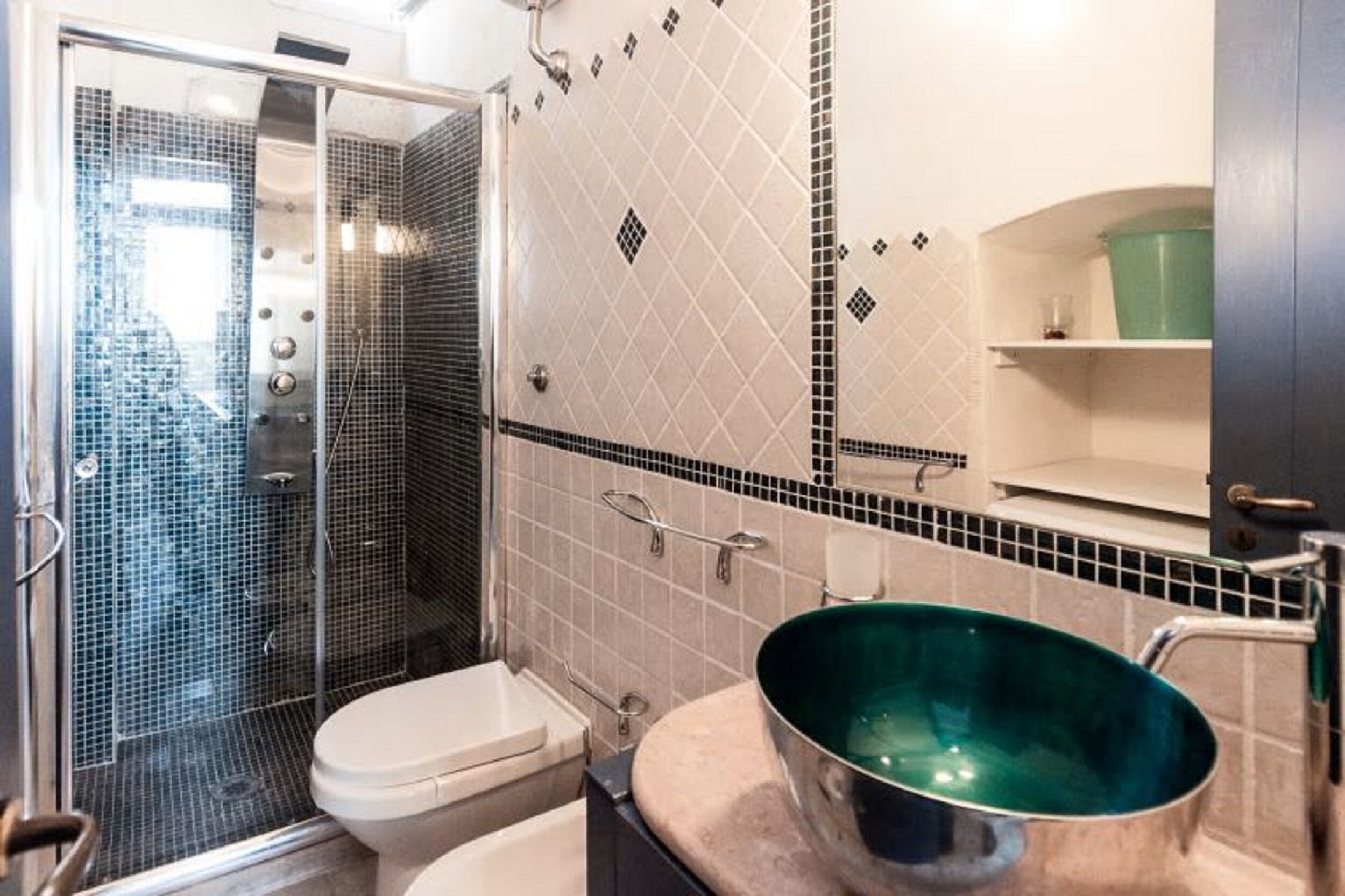 Ein Badezimmer mit Duschkabine aus Glas, Toilette, Waschbecken mit grüner Schüssel und Fliesenwand mit Rautenmuster.