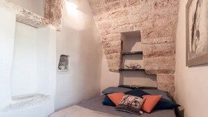 A bedroom with a brick wall, a bed with pillows, and a small shelf. - Biancocalce (Ostuni)