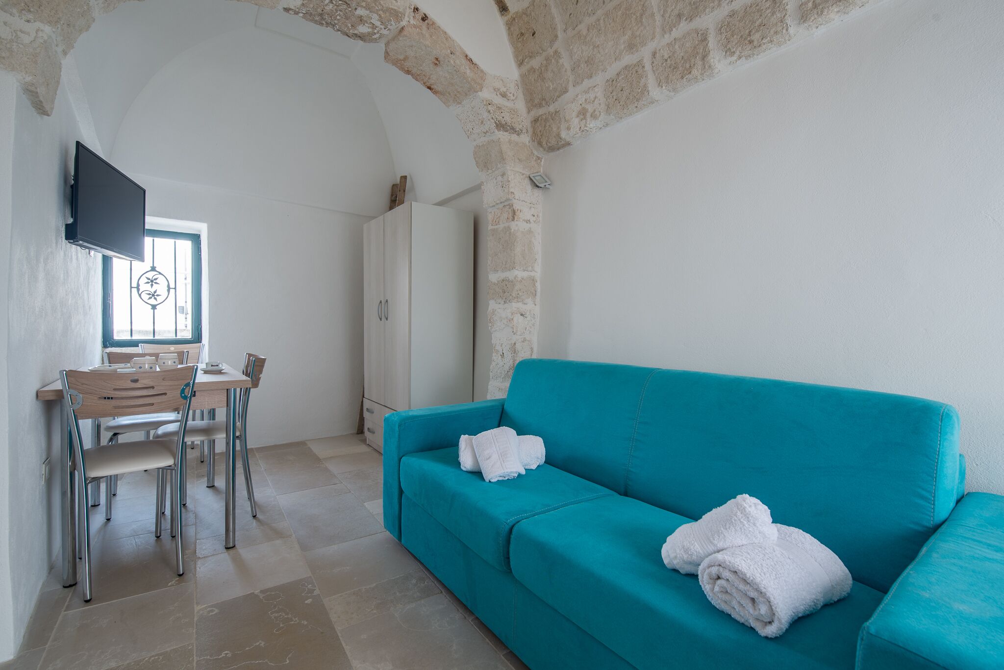 Una habitación con un sofá color turquesa, una mesa de comedor con sillas, un televisor montado en la pared y un ventanal con un diseño decorativo.