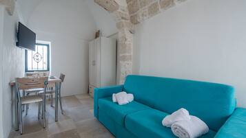 Una habitación con un sofá color turquesa, una mesa de comedor con sillas, un televisor montado en la pared y un ventanal con un diseño decorativo.