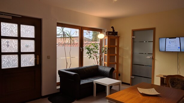 Una sala de estar con un sofá negro, una mesa de centro blanca, un tapete estampado, una mesa de comedor de madera y un televisor montado en la pared.