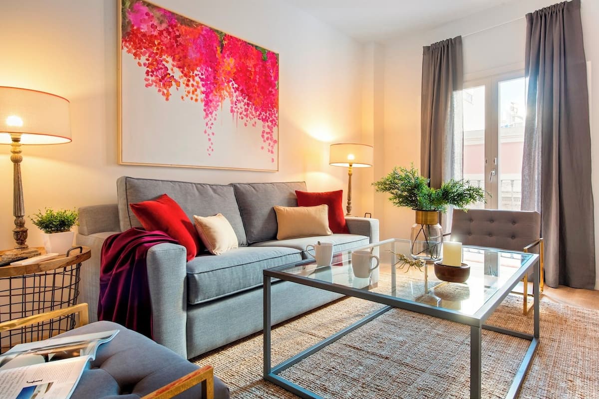 Un salon moderne avec un canapé gris, des coussins rouges et beige, une table basse en verre et un grand tableau abstrait accroché au mur.