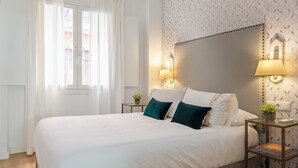 A neatly made bed with white linens and two teal pillows, a bedside table with a lamp, and a wall-mounted light fixture. - Genteel Home Baños (Seville)