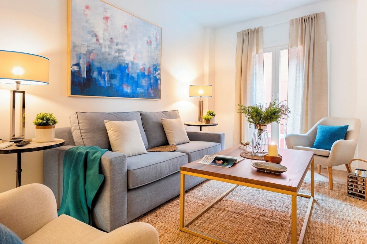 Un salon moderne avec un canapé gris, une table basse et un fauteuil bleu. On y trouve deux lampes sur des tables d’appoint, un tableau au mur et une fenêtre avec des rideaux.