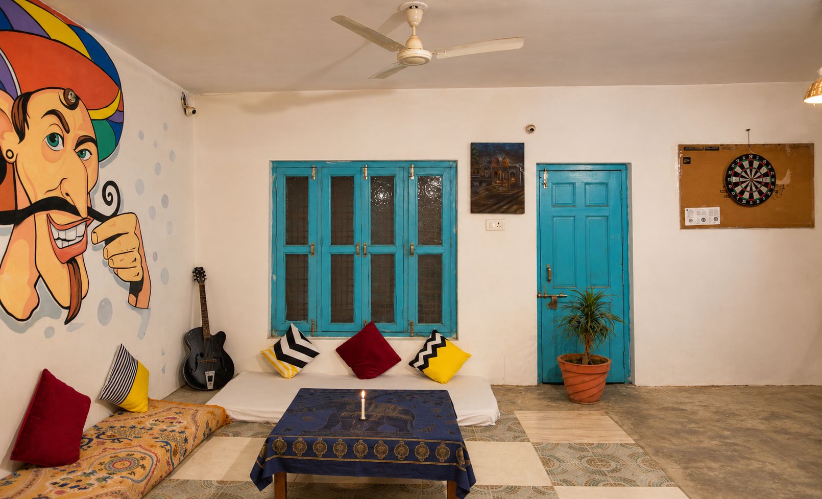 A room with a bed, a guitar, a table with a tablecloth, a ceiling fan, and a door with a dartboard.