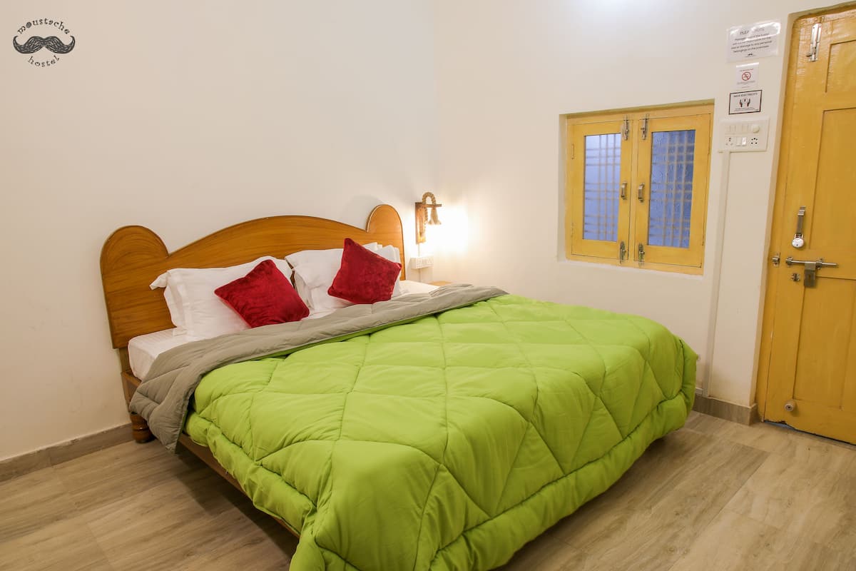 A neatly made bed with a green quilt, a wooden headboard, and red pillows. There is a wall-mounted light fixture above the bed and a window with a wooden frame.