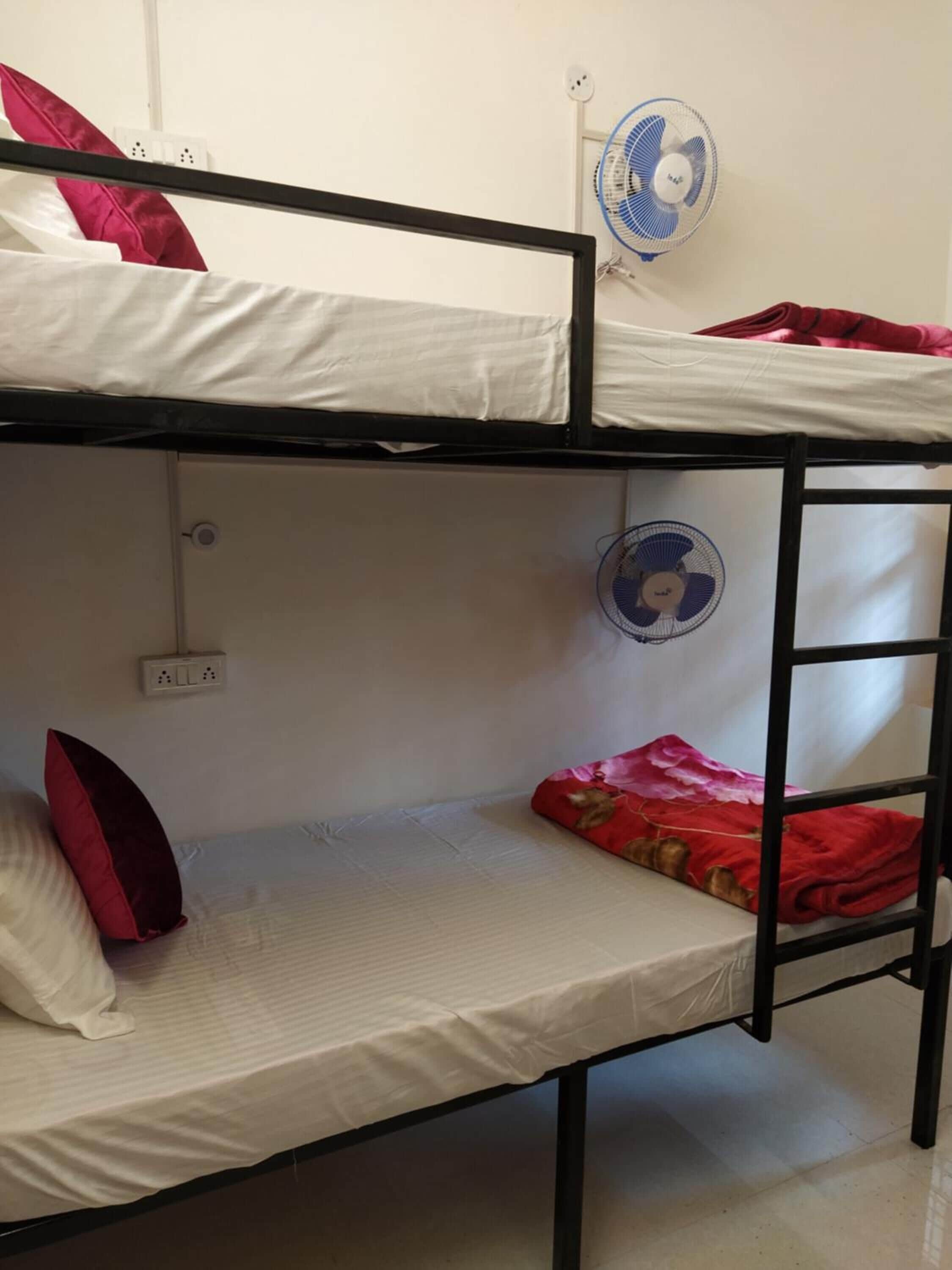 Habitación con literas, dos ventiladores, un enchufe y una almohada roja en la litera inferior.