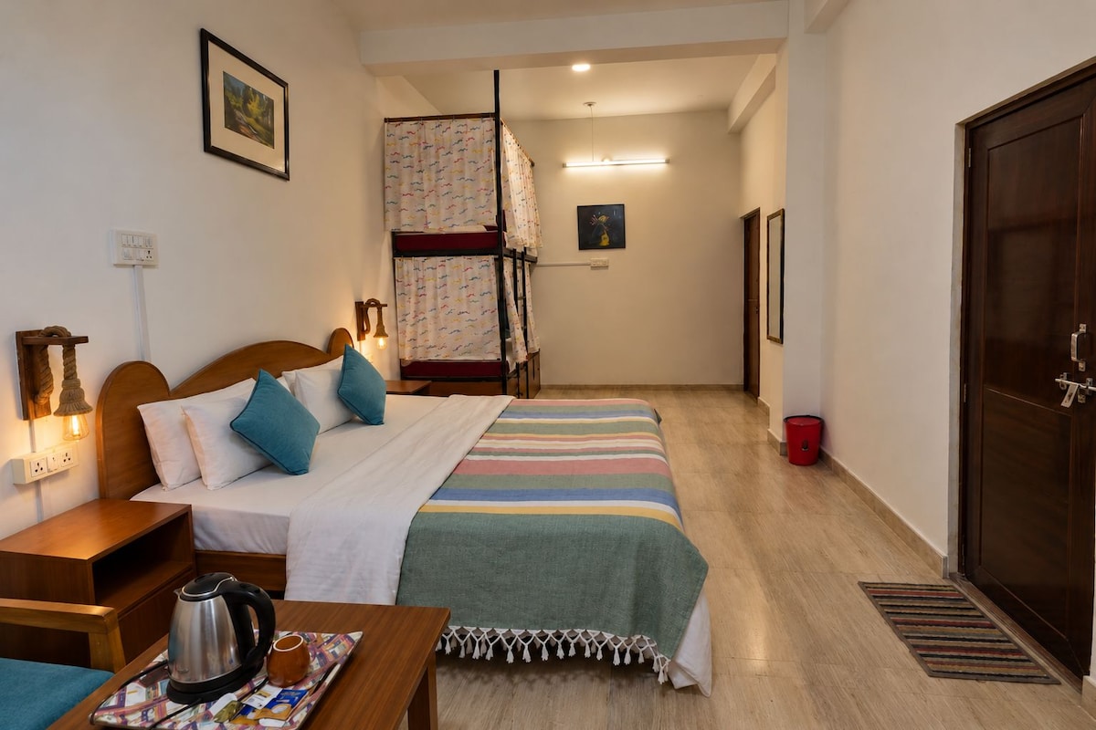 A hotel room with a bed, a wooden nightstand, a table with a kettle and a magazine, a bunk bed, and a door.