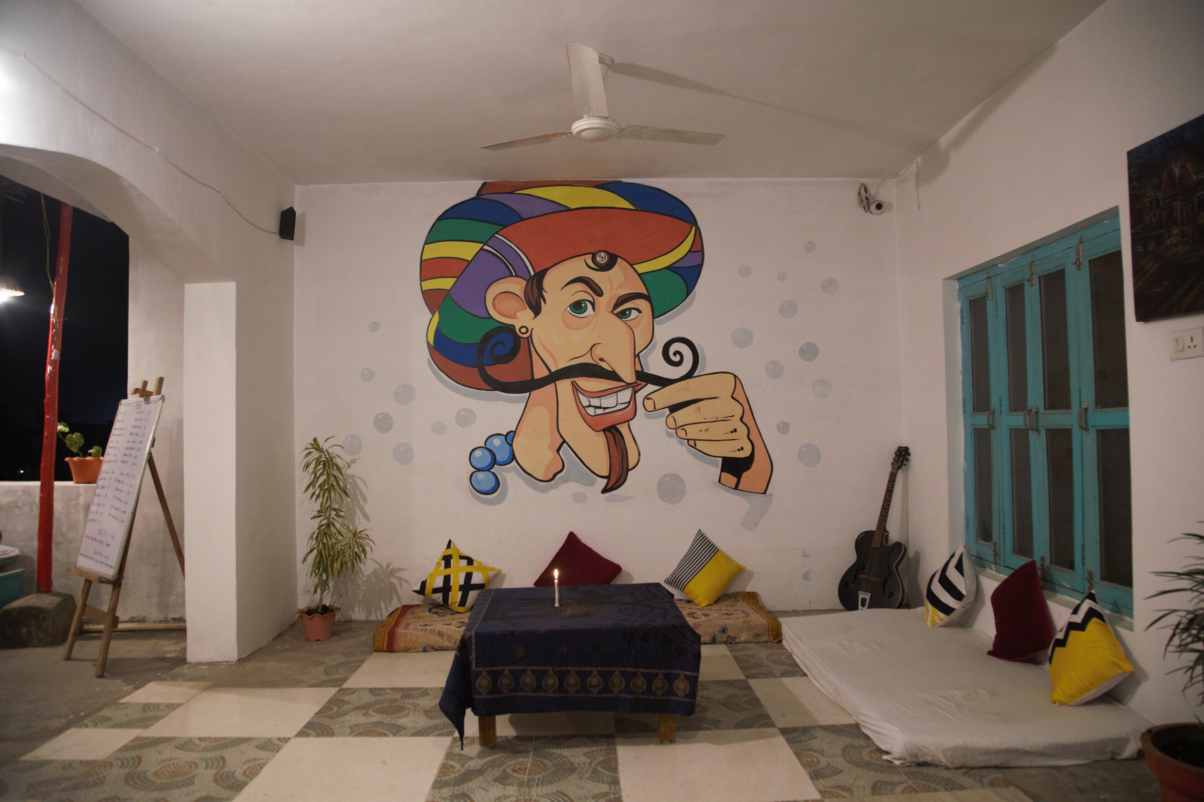 Una habitación con un mural grande, un ventilador de techo, una guitarra y una mesa con una vela.