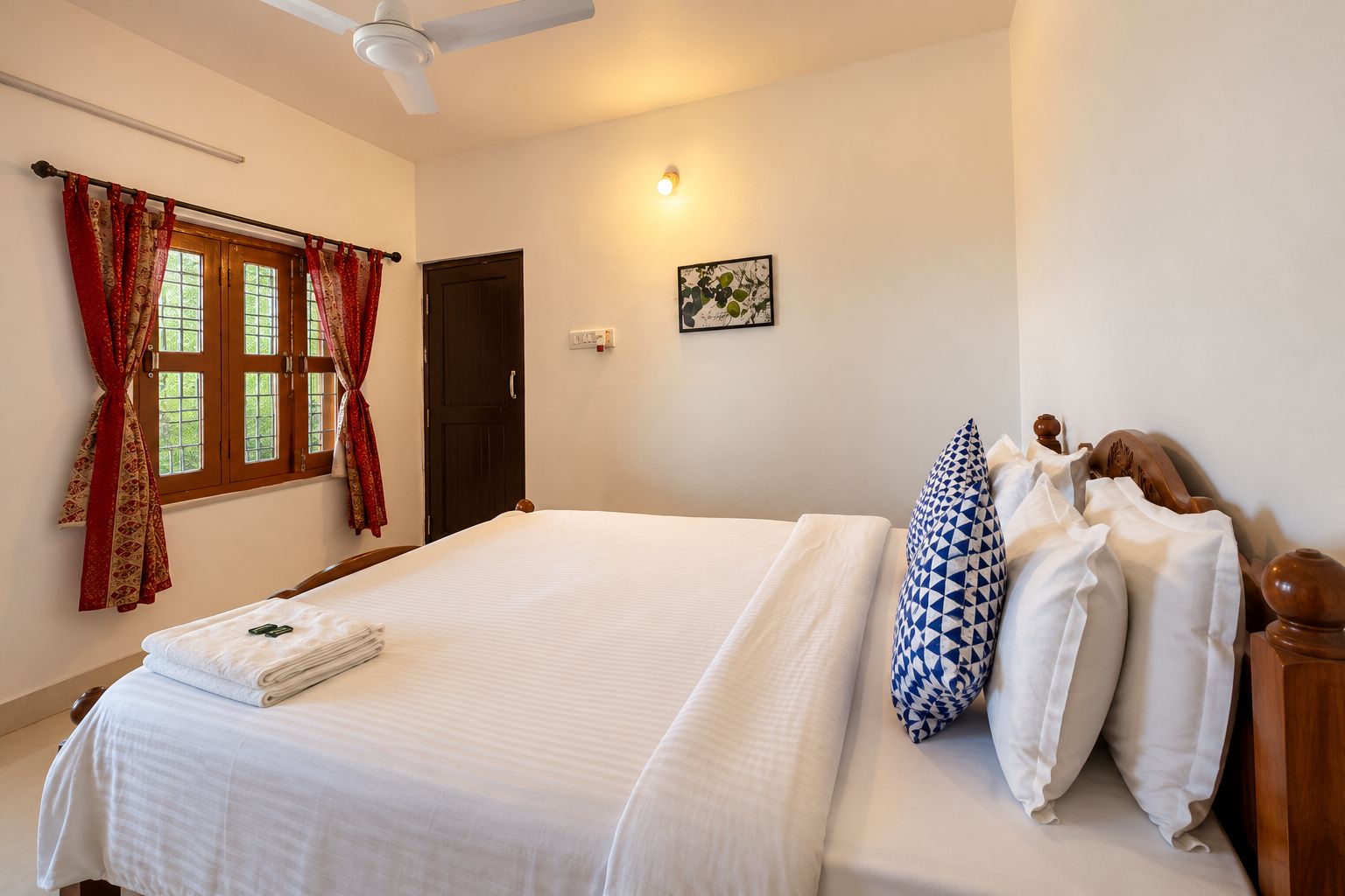 A hotel room with a bed, a ceiling fan, a window with curtains, a door, and a wall-mounted light.