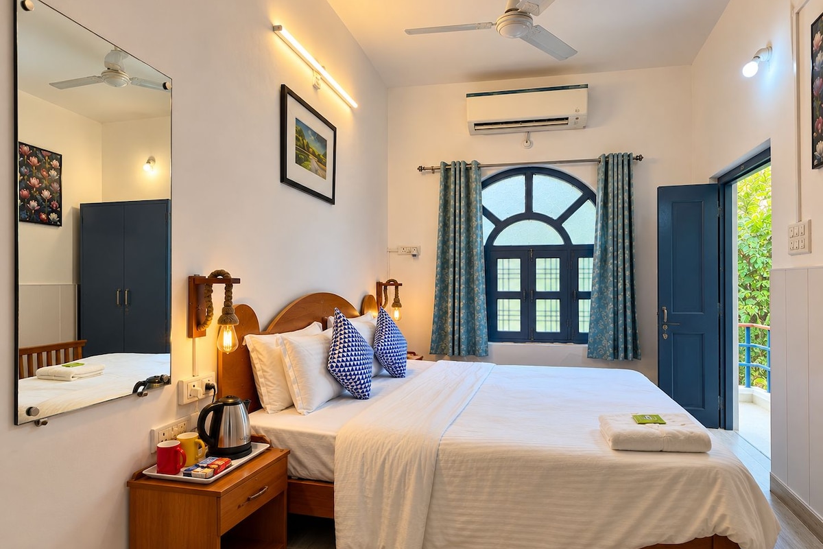 A hotel room with a bed, bedside table, mirror, window with curtains, and an air conditioning unit.