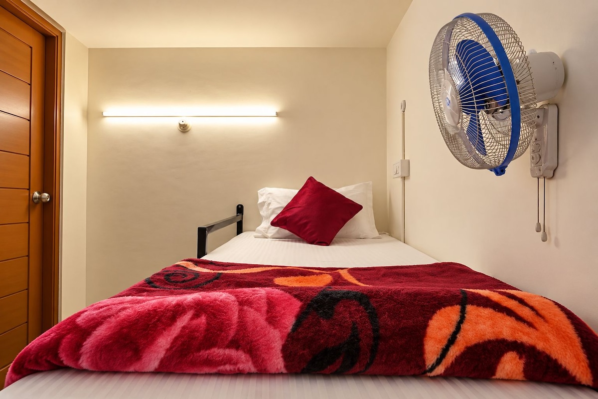 A single bed with a red pillow, a patterned blanket, and a wall-mounted fan.