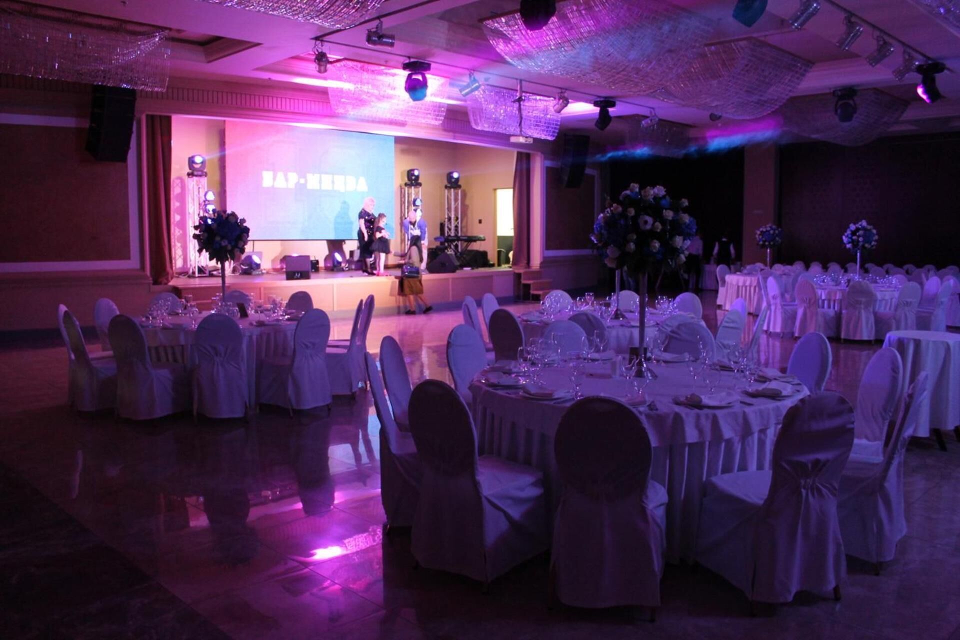 A banquet hall with round tables set for dining, a stage with a presentation screen, and a chandelier.
