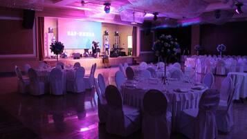 A banquet hall with round tables set for dining, a stage with a presentation screen, and a chandelier.