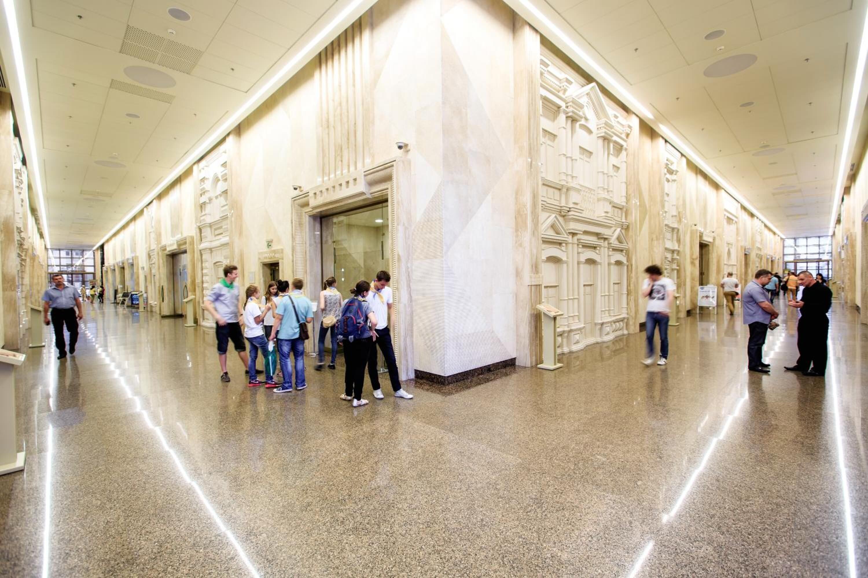A spacious corridor with marble walls and a polished floor, lined with doors and a few people walking.