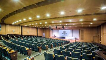 Un gran auditorio con filas de sillas y un escenario con un atril y una pantalla que muestra un logotipo.
