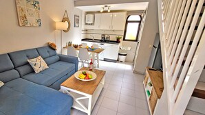A living room with a blue sofa, a wooden coffee table with fruit, and a kitchen in the background.