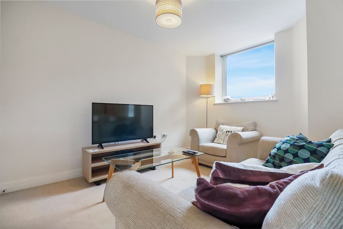 A modern living room with a flat-screen TV, a glass coffee table, a sofa, a lamp, and a window with a view.