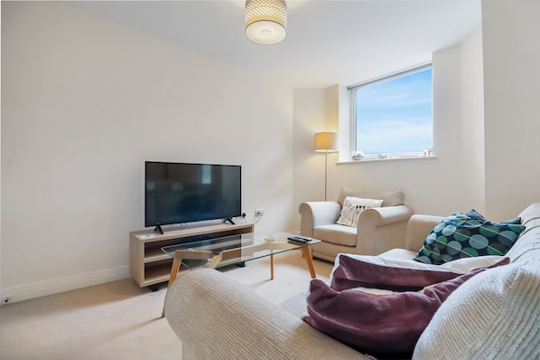 A modern living room with a flat-screen TV, a glass coffee table, a sofa, a lamp, and a window with a view. - Stevenage Apartments (Stevenage)