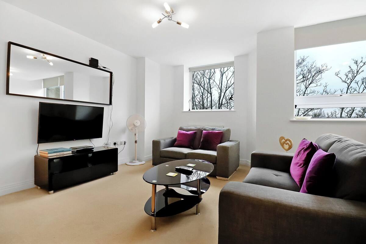 A modern living room with a flat-screen TV, a coffee table, two sofas, and a large window with a view of bare trees.