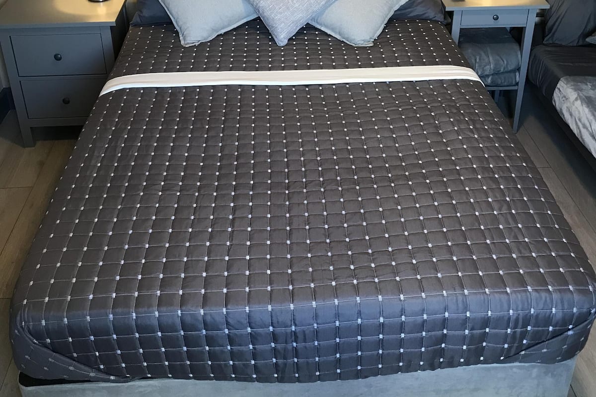 Una cama bien hecha con cubrecolchón acolchado, dos mesitas de noche con lámparas, una obra de arte en la pared y una cama nido a la derecha.