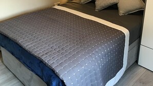 Ein ordentlich bezogenes Bett mit grauer Decke und Kissen, einer Nachttischlampe und einem Holzboden.