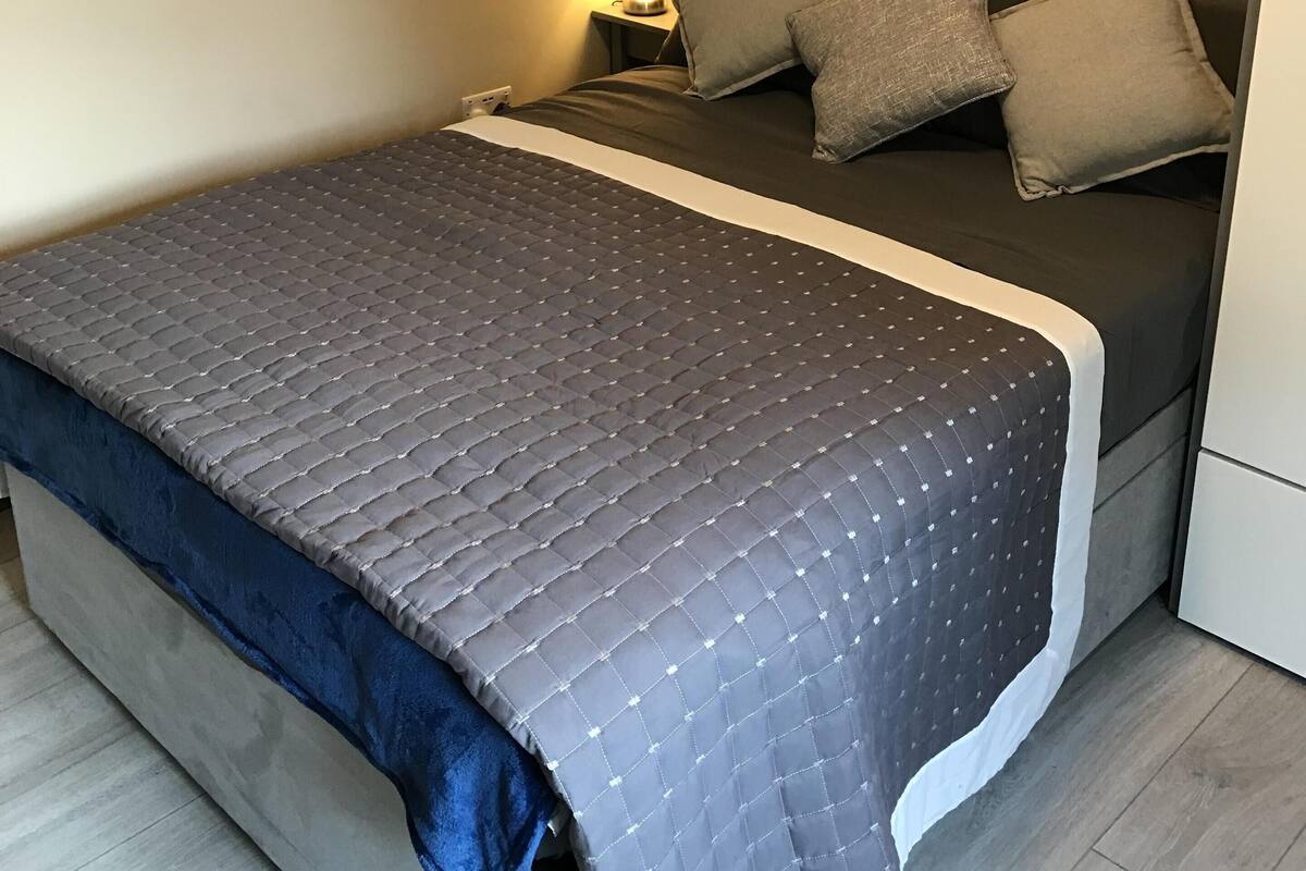 Una cama bien hecha con colcha oscura, dos almohadas y una lámpara de noche.