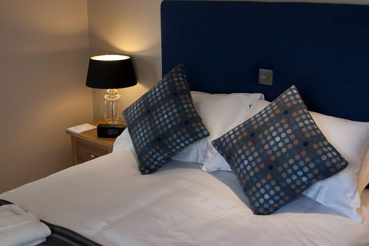 A neatly made bed with white linens and two blue pillows with a polka dot pattern. There is a lamp on a bedside table to the left, and a painting of a sunset on the wall above the bed.