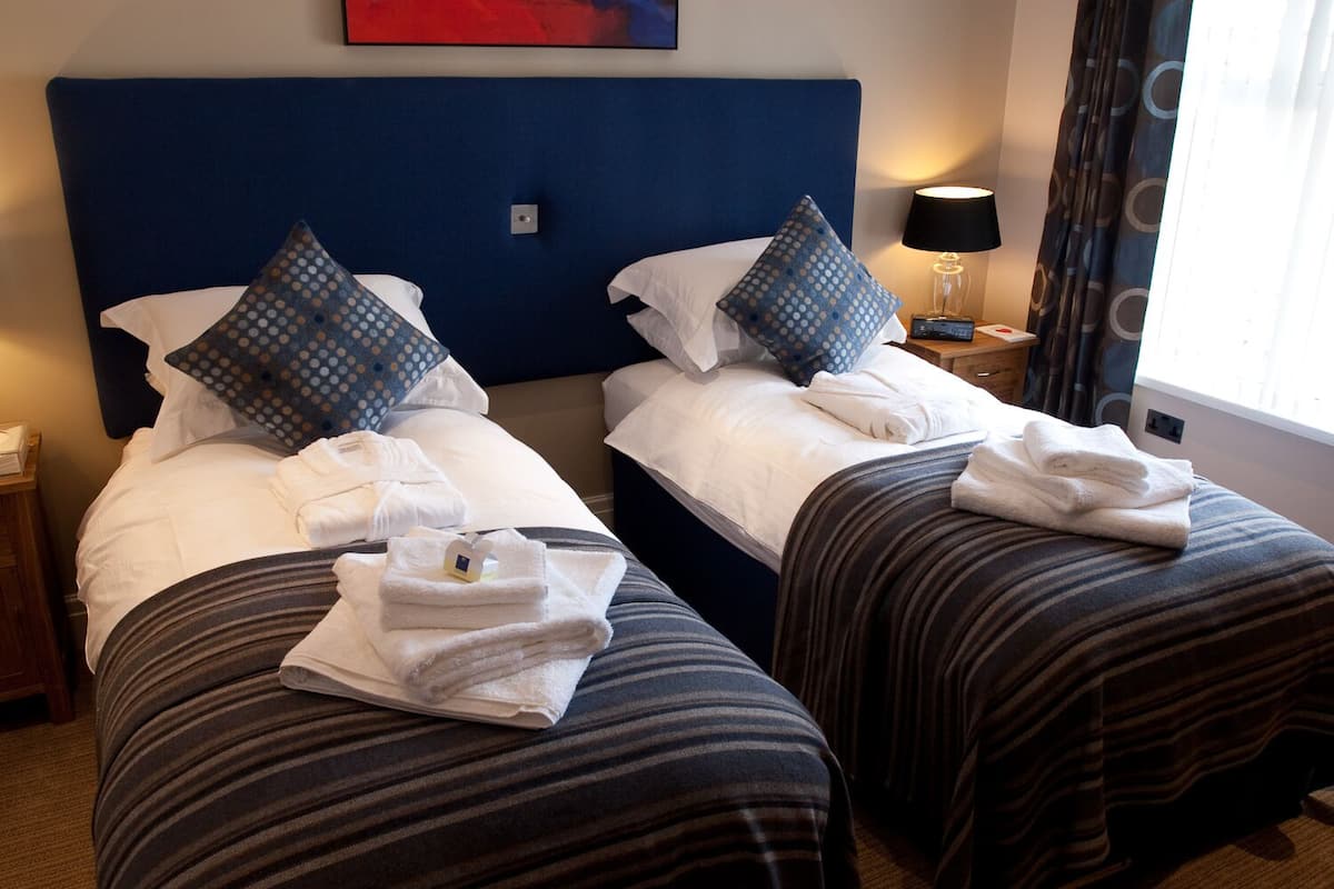 A hotel room with two beds, each with white linens and blue polka dot pillows. There are folded towels on the beds and a nightstand with a lamp and a clock.