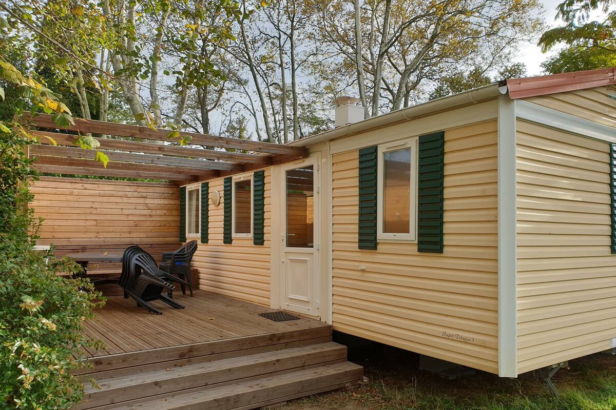 Une maison mobile de plain-pied dotée d’une terrasse en bois, d’un patio couvert et d’un petit espace extérieur aménagé avec deux chaises.