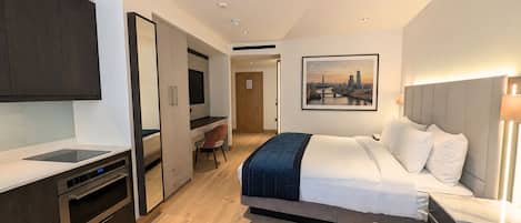 A modern hotel room with a bed, desk, and kitchenette.