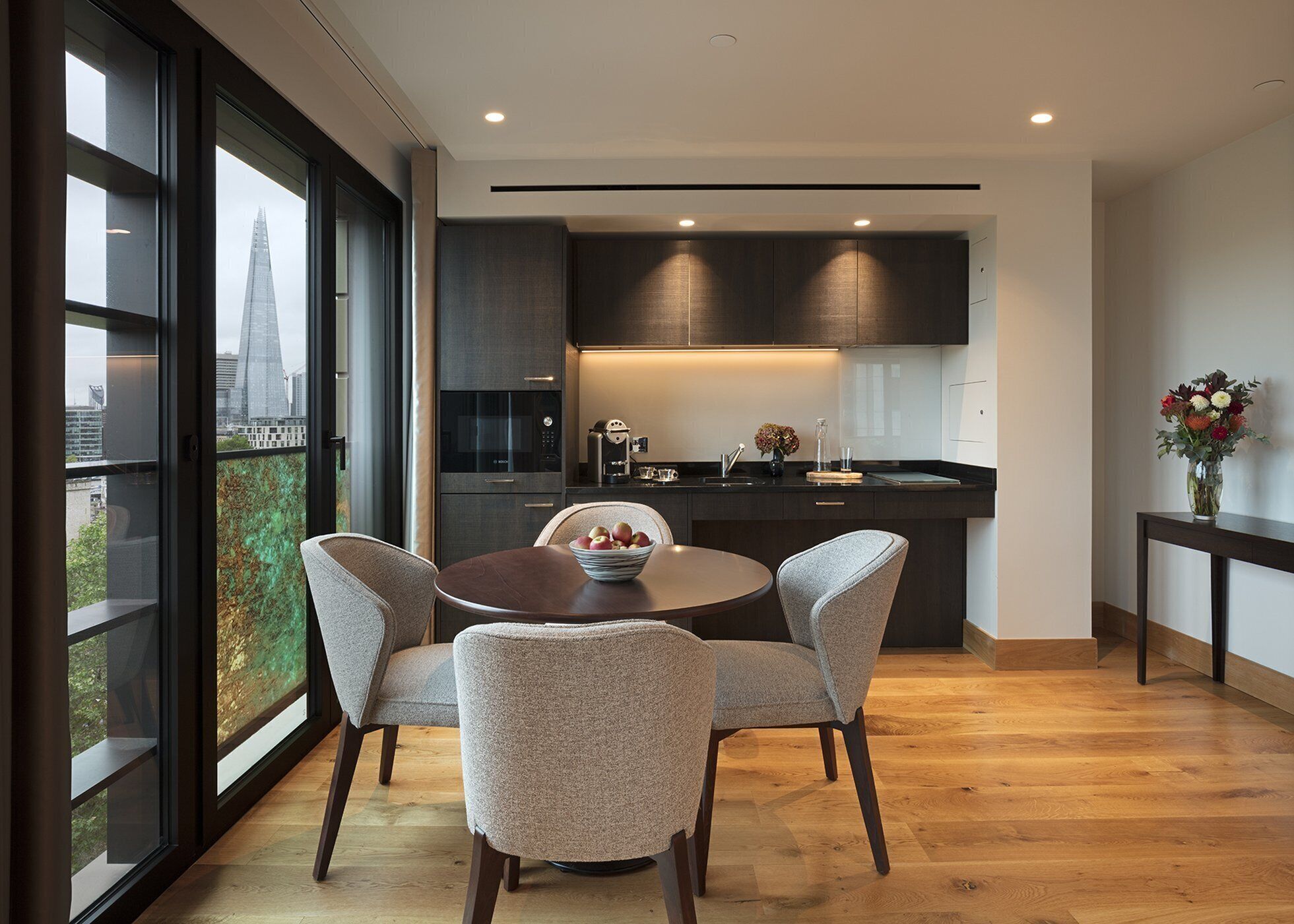 A modern kitchen with dark cabinetry, a round dining table with chairs, and a view of a cityscape through large windows.