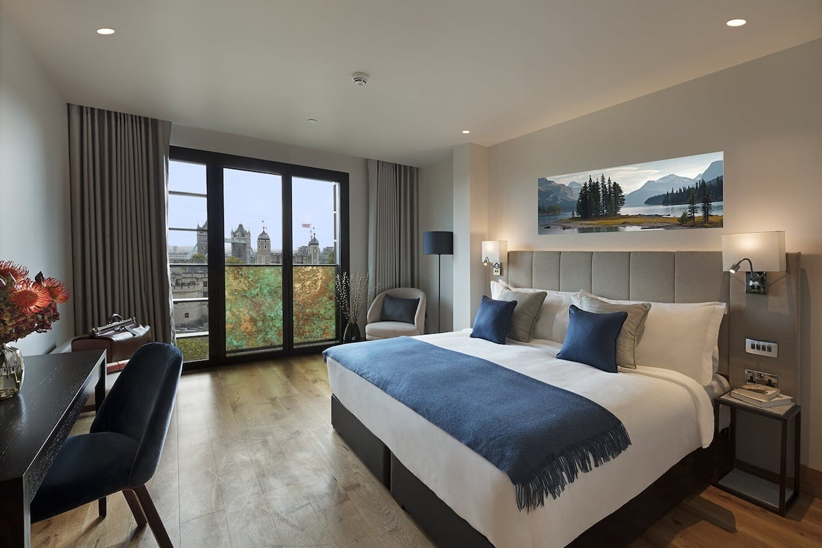 A modern hotel room with a large bed, a desk, and a chair. There is a view of a cityscape through the window.