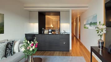 Ein modernes Wohnzimmer mit einer Couch, einem Couchtisch und einer Blumenvase. Im Hintergrund ist eine Küchenzeile mit Arbeitsplatte und Schränken erkennbar.
