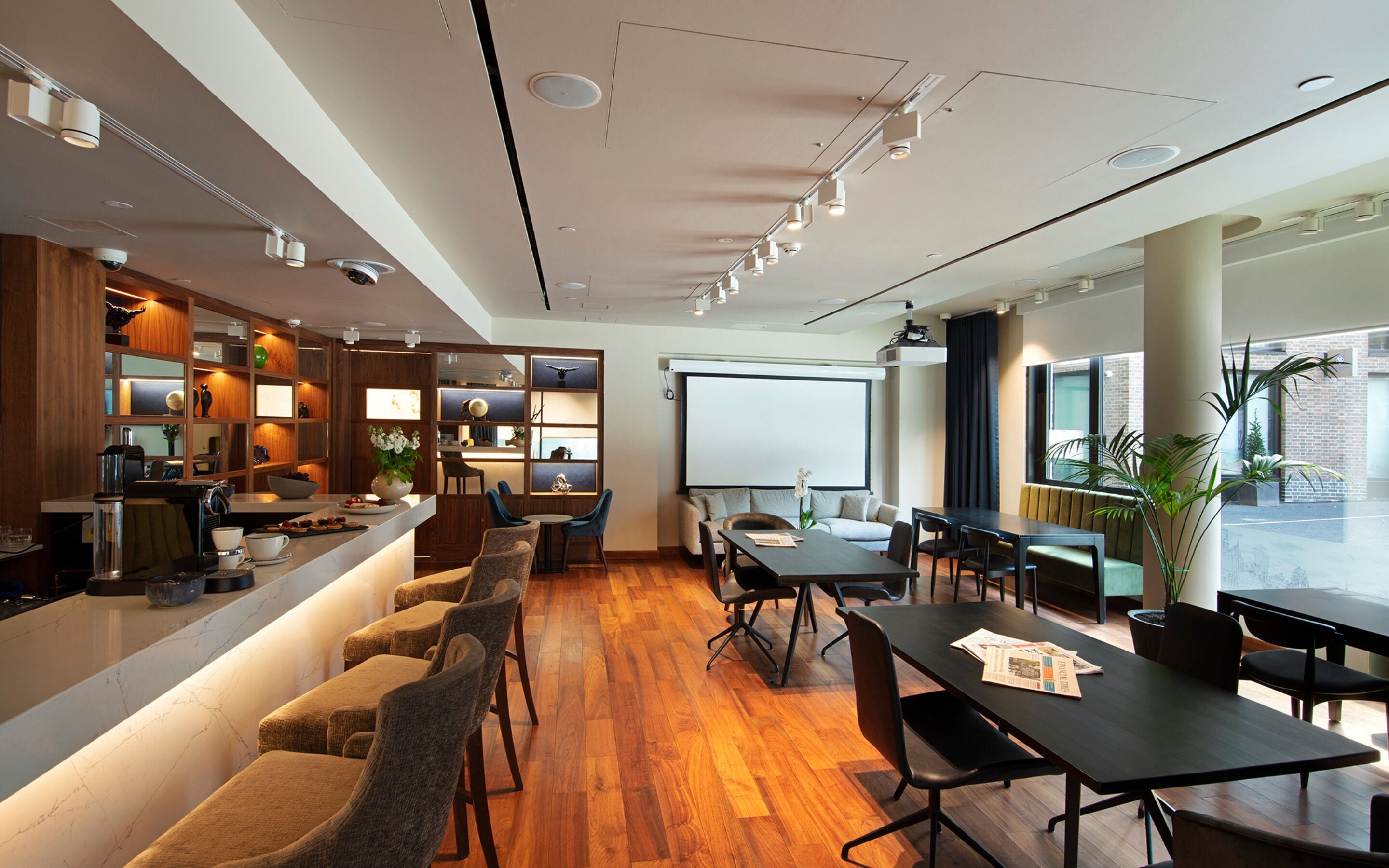 A modern conference room with a long table, several chairs, a projector screen, and a coffee station with a counter and seating area.