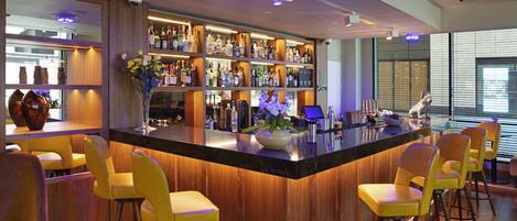 A modern bar area with a wooden bar counter, yellow bar stools, and a variety of bottles and glasses.