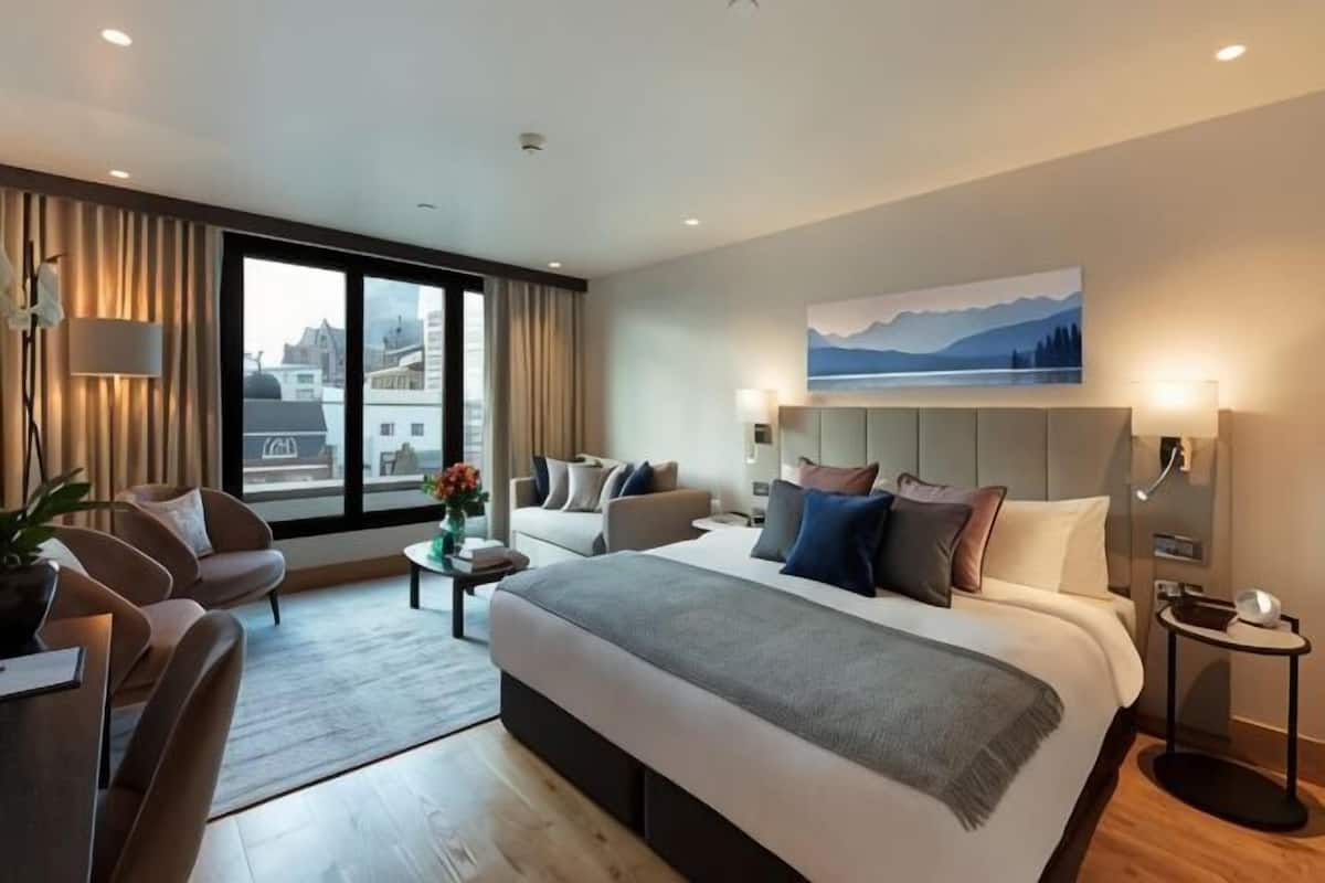 A modern hotel room with a large bed, a sofa, a desk, and a view of the city through large windows.
