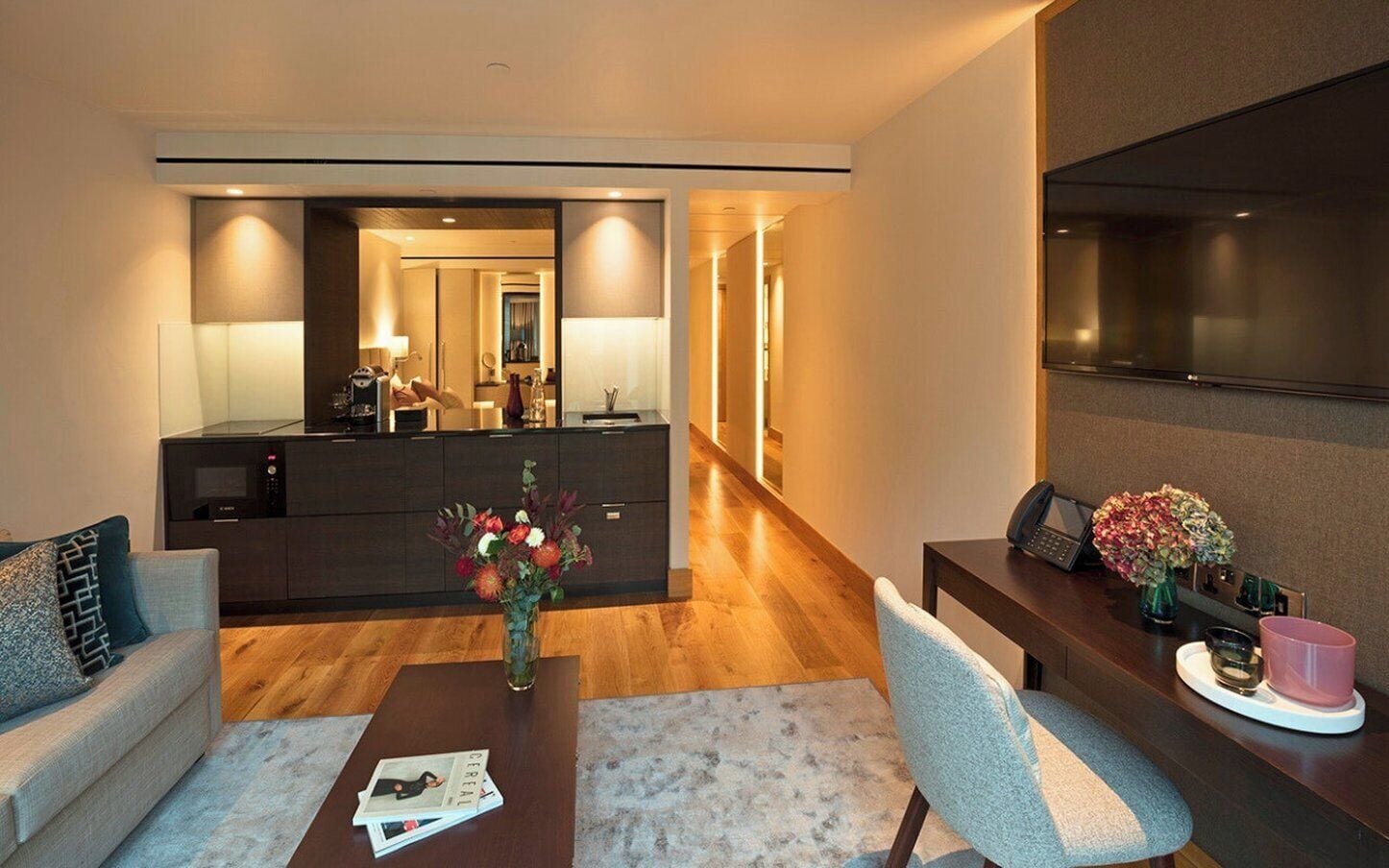 A modern hotel room with a living area, a dining table, a kitchen area, and a large flat-screen TV.
