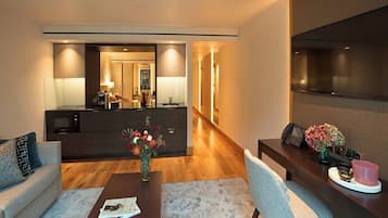 A modern hotel room with a living area, a dining table, a kitchen area, and a large flat-screen TV.