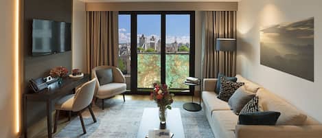 Un salon moderne comprenant un canapé, un fauteuil, une table basse et un bureau avec un ordinateur. Une grande fenêtre offre une vue sur la ville.