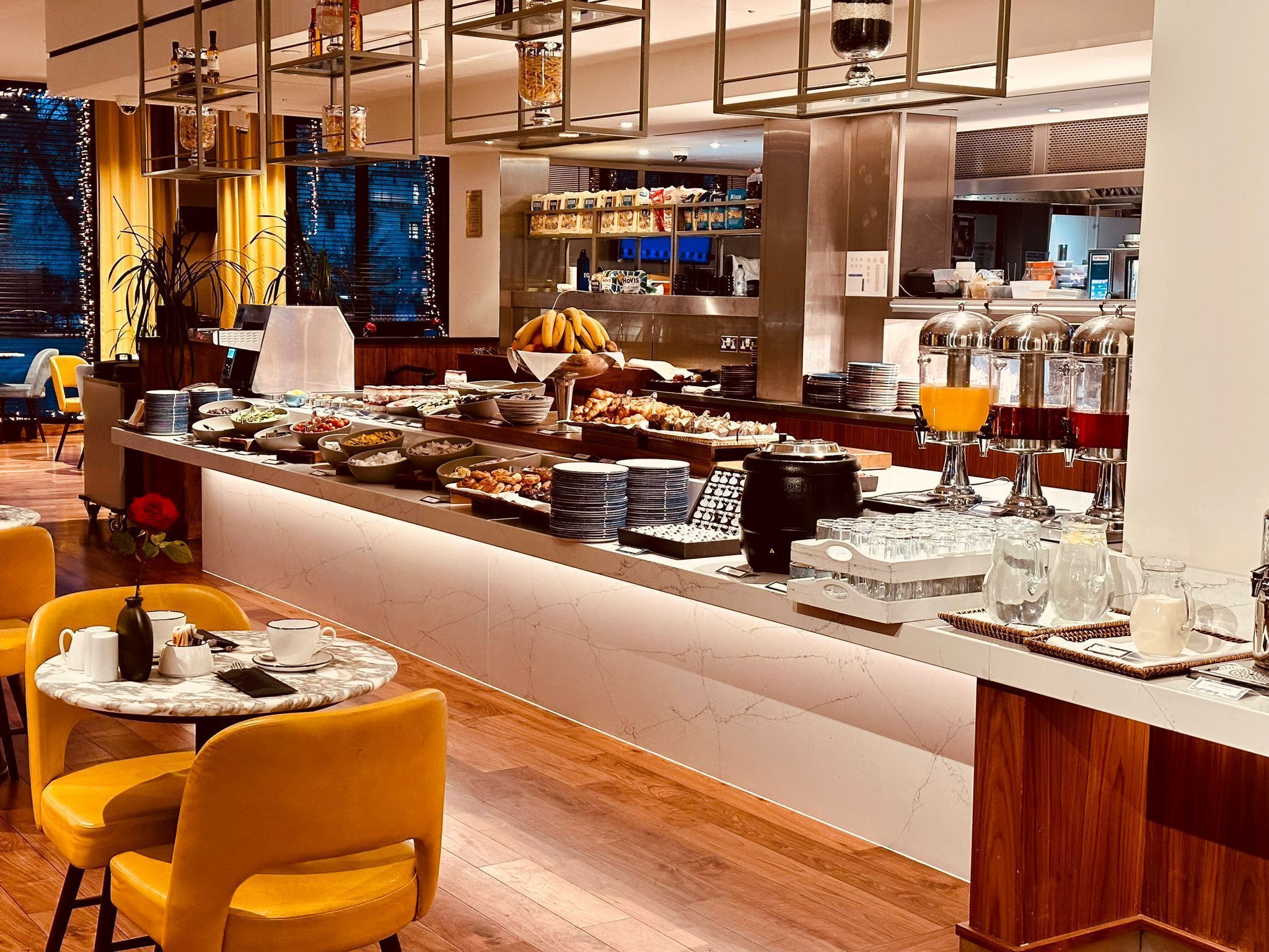 A breakfast buffet with various food items, a coffee station, and seating area with yellow chairs.