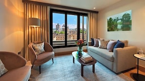 A modern living room with a sofa, armchair, and coffee table, a large window with a city view, and a painting on the wall. - Tower Suites by Blue Orchid (London)