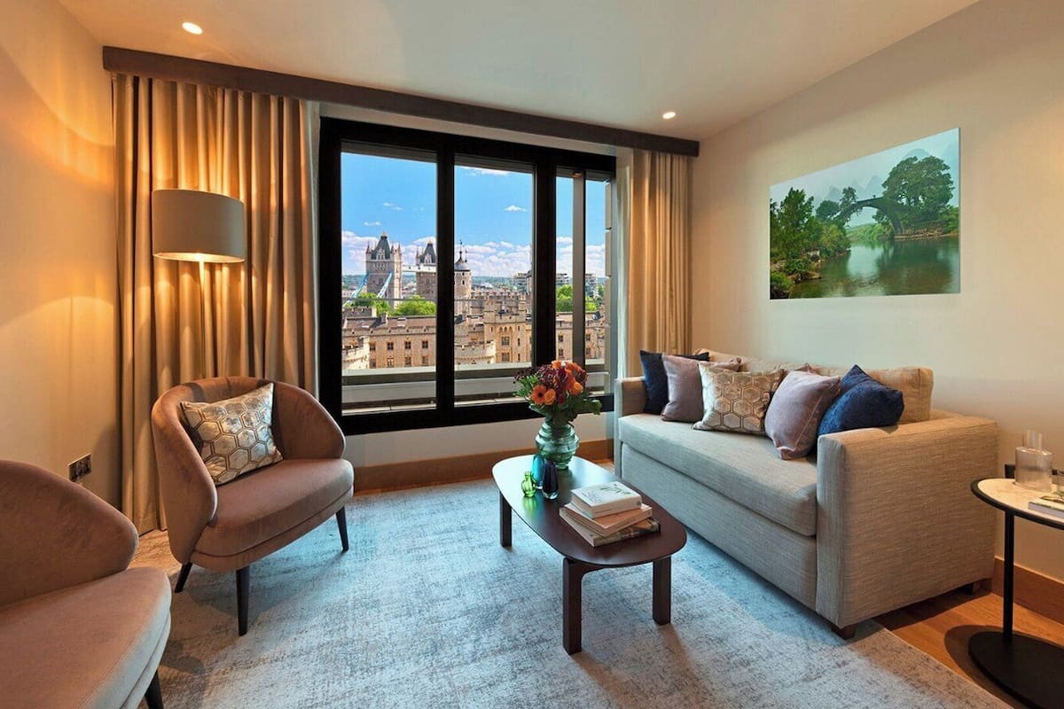 A modern living room with a sofa, armchair, and coffee table, a large window with a city view, and a painting on the wall.