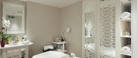 Une salle de spa moderne équipée d’une table de massage, d’une coiffeuse avec un miroir et d’étagères où sont rangées les serviettes.
