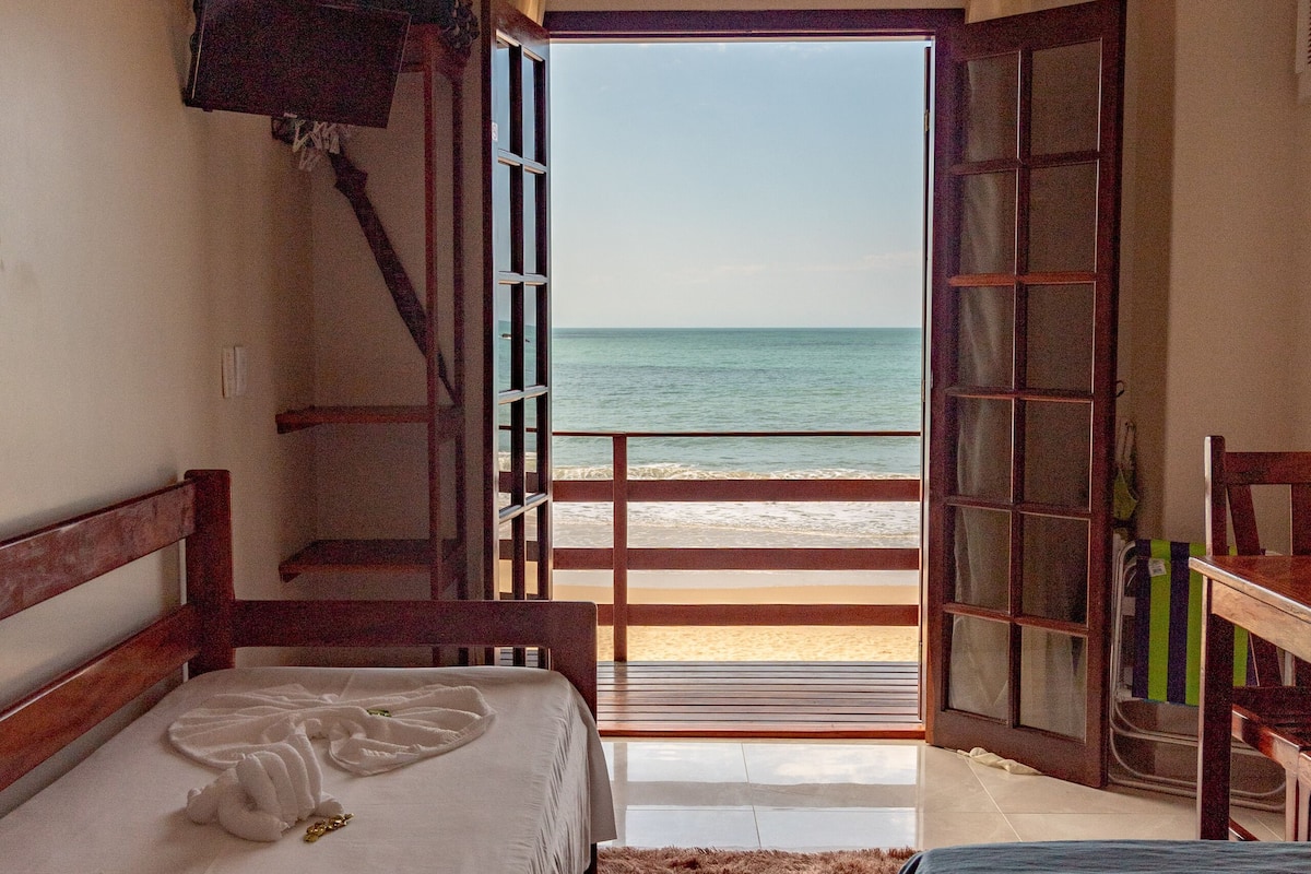 Ein Zimmer mit einem Bett, einem Sessel, einem kleinen Tisch, einem Deckenventilator und Blick auf den Strand.