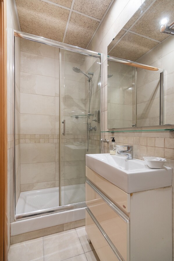 A modern bathroom with a glass-enclosed shower, a white sink with a mirror above it, and beige tiles on the walls and floor. - Hostal House (Barcelona)