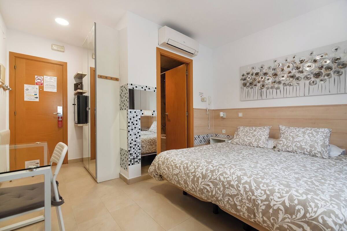 Quarto de hotel com cama, escrivaninha e cadeira. Porta com espelho e telefone fixado na parede.
