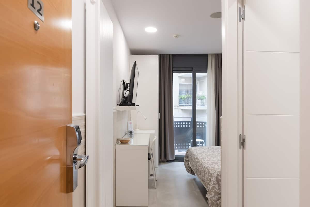 Quarto de hotel com porta de madeira, guarda-roupa branco, pequena cozinha e cama com roupa de cama estampada, além de vista para a varanda.