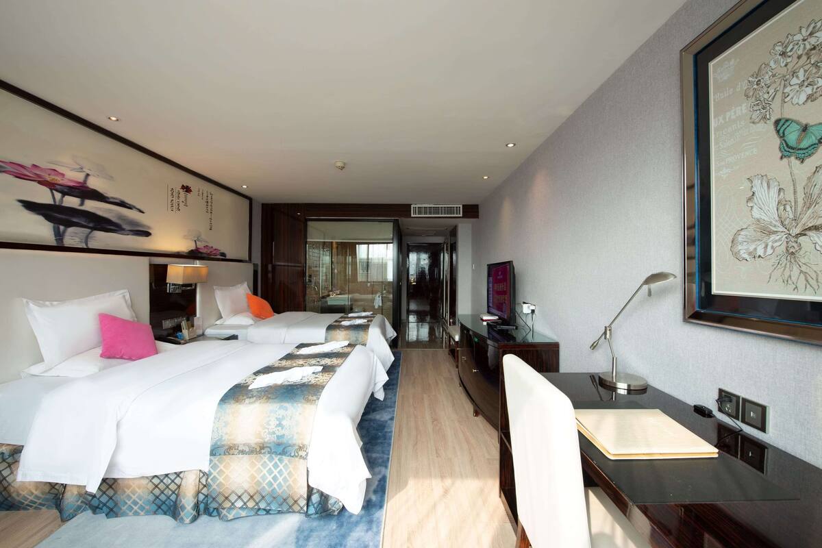 Habitación de hotel con dos camas, un escritorio, una silla, un televisor y un ventanal grande.