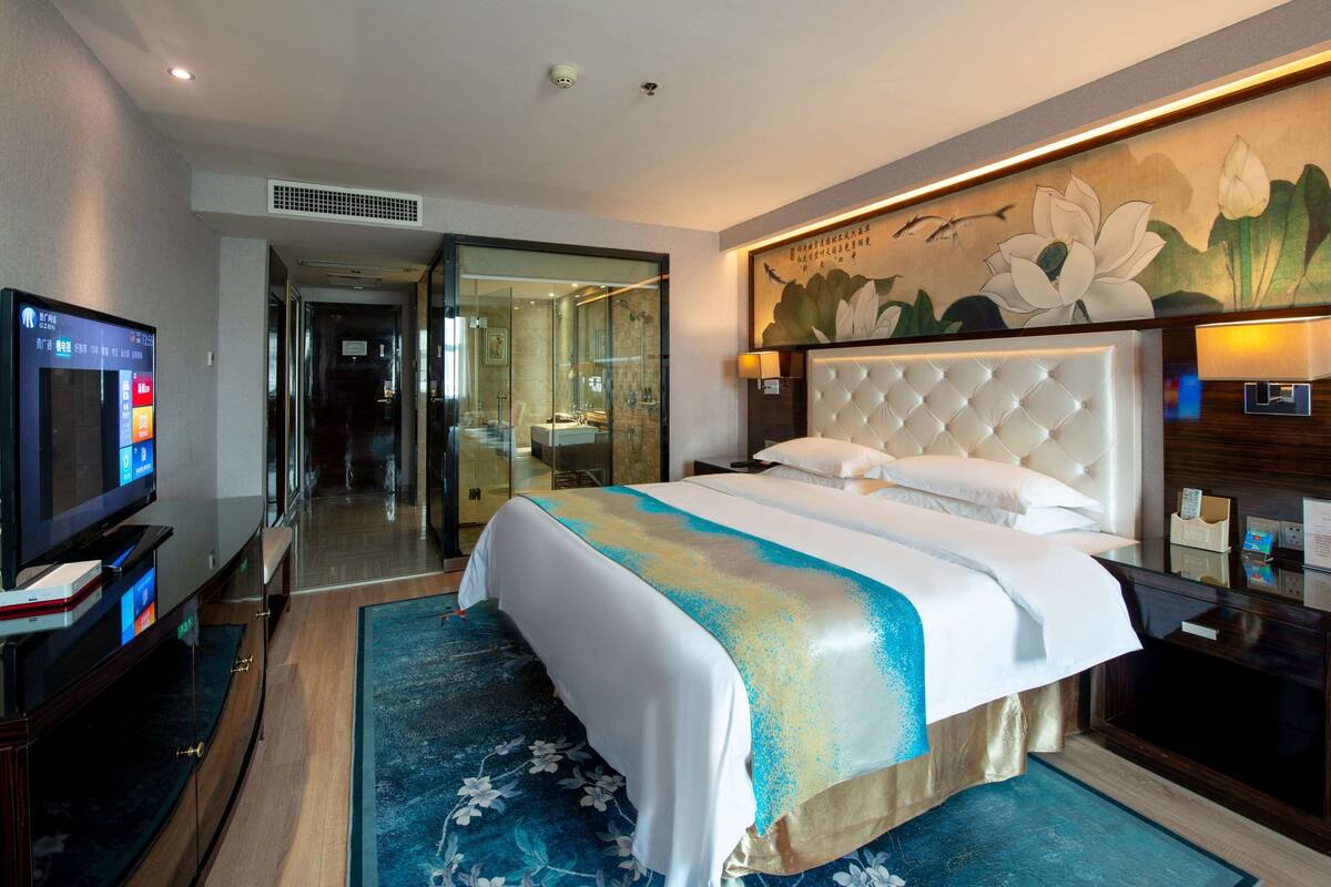 Una habitación de hotel con una cama grande, un televisor, un baño con puerta de vidrio y un cuadro en la pared.